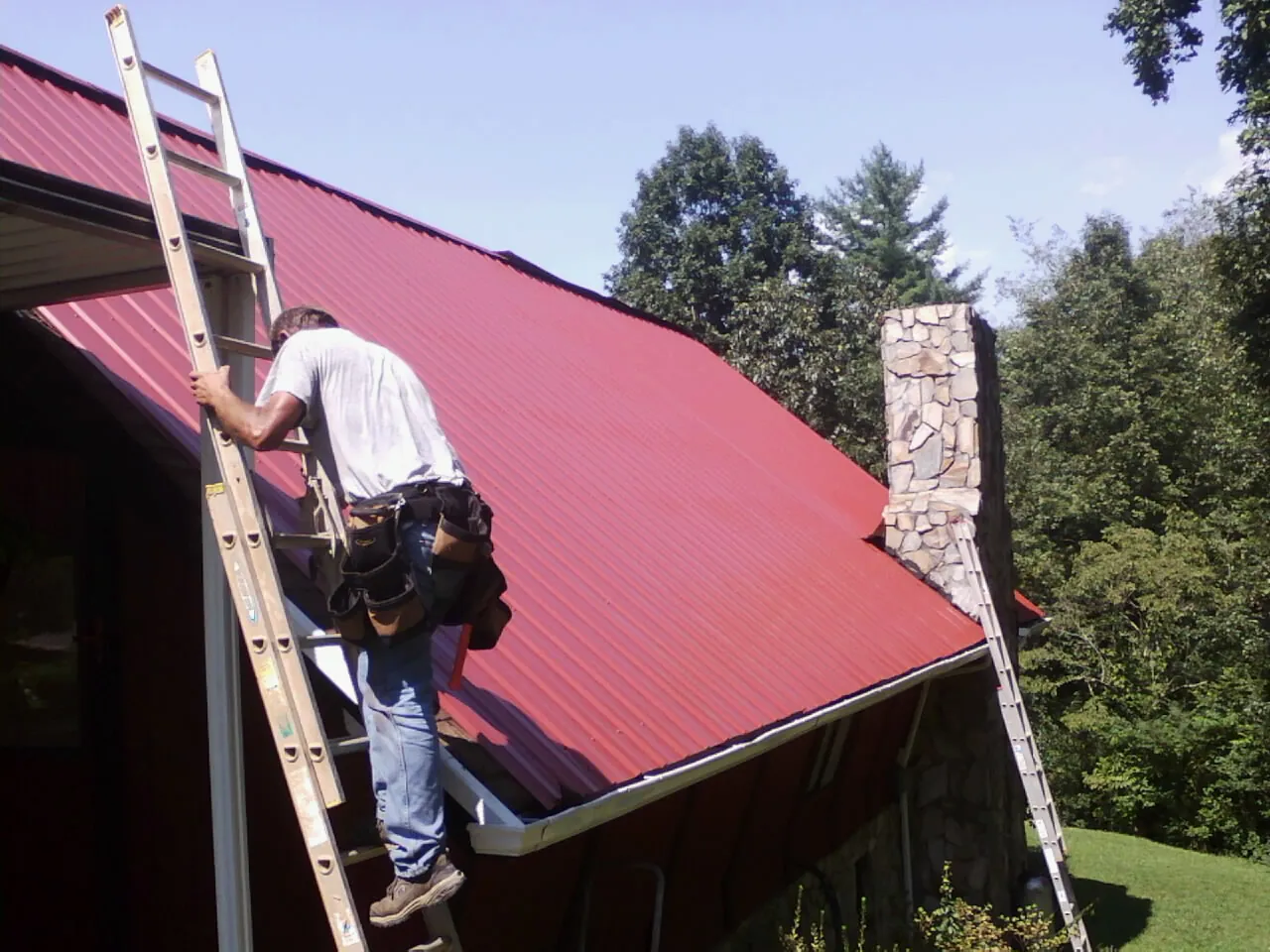 Roofer on a ladder beside a red metal roof and stone chimney — Soffit & Fascia Repair by Roofing Brothers in Port Orford